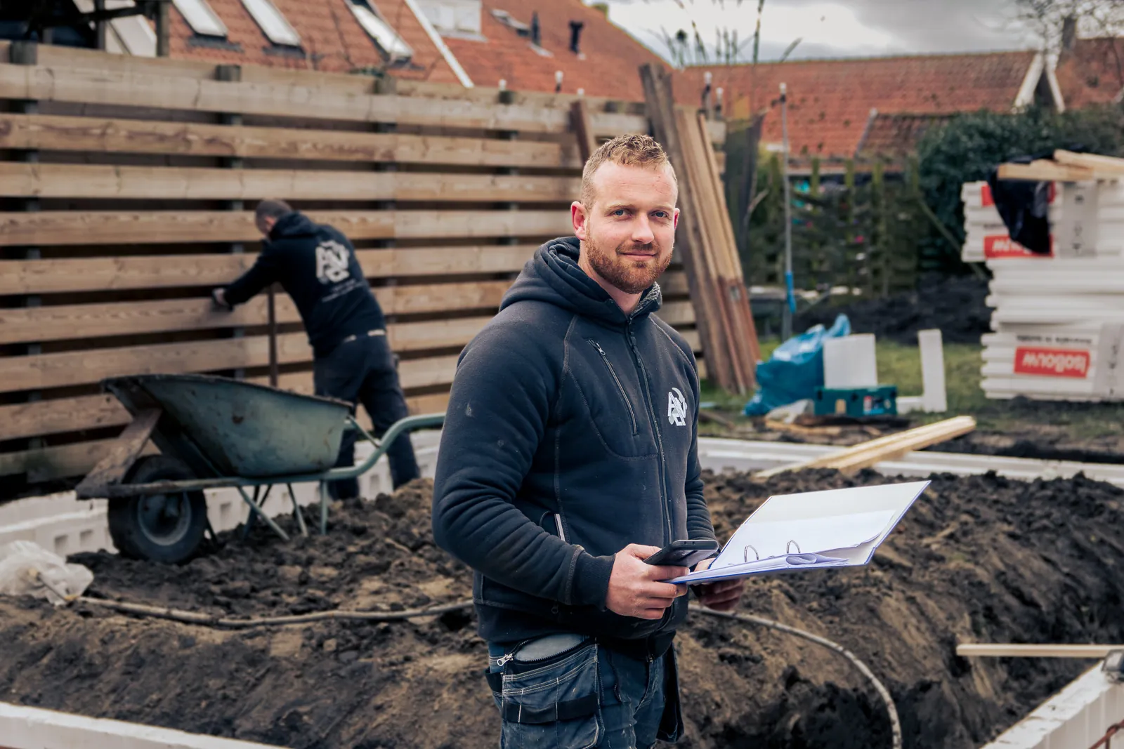 Een vakman van ADV met de bouwtekening in de hand; achter hem wordt de grond voor een fundering voorbereid.