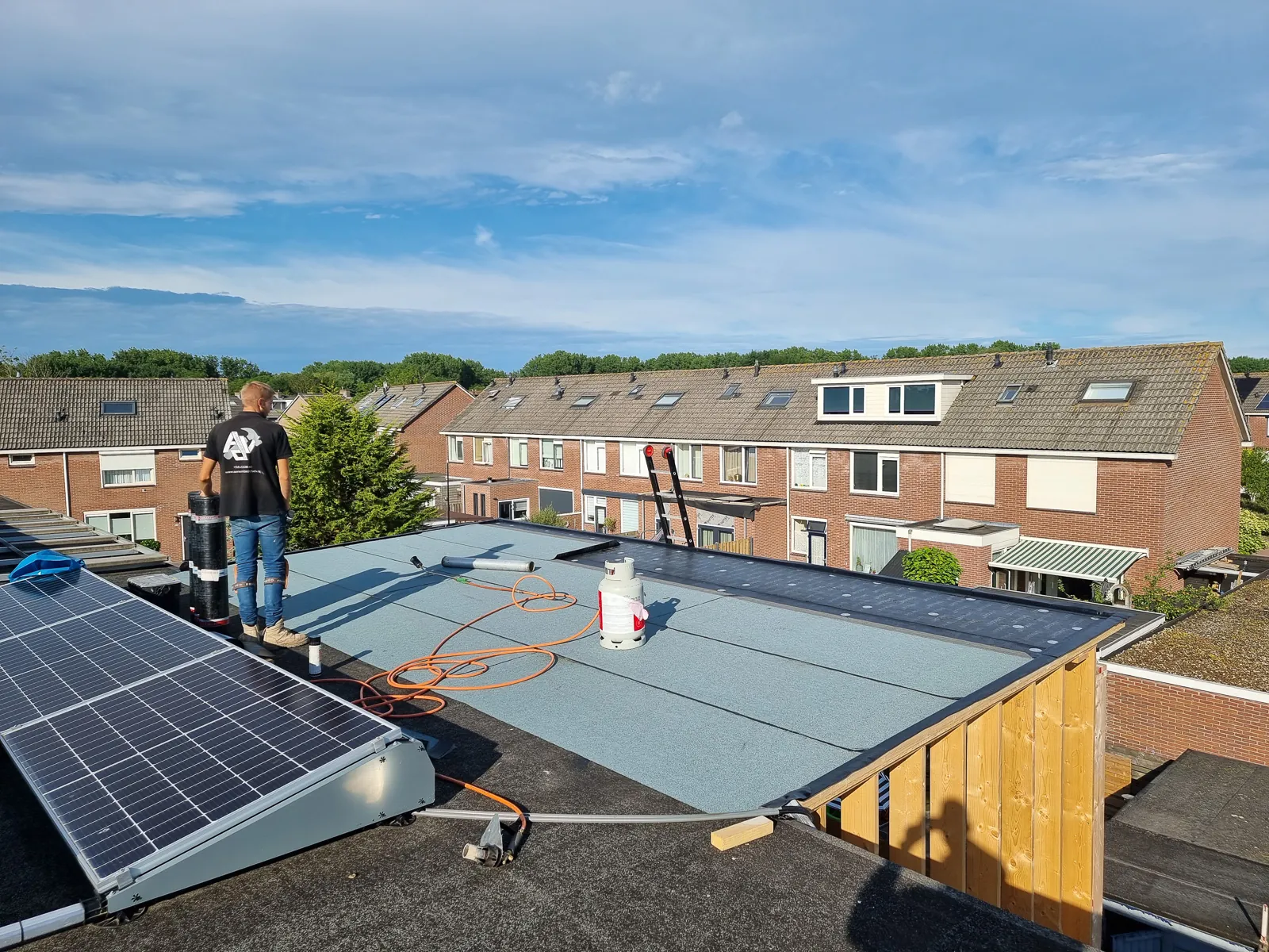 Een vakman op een plat dak met zonnepanelen boven een rij woningen.